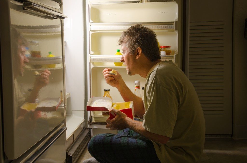man eating as he sits in front of a refrigerator eating ice cream out of carton